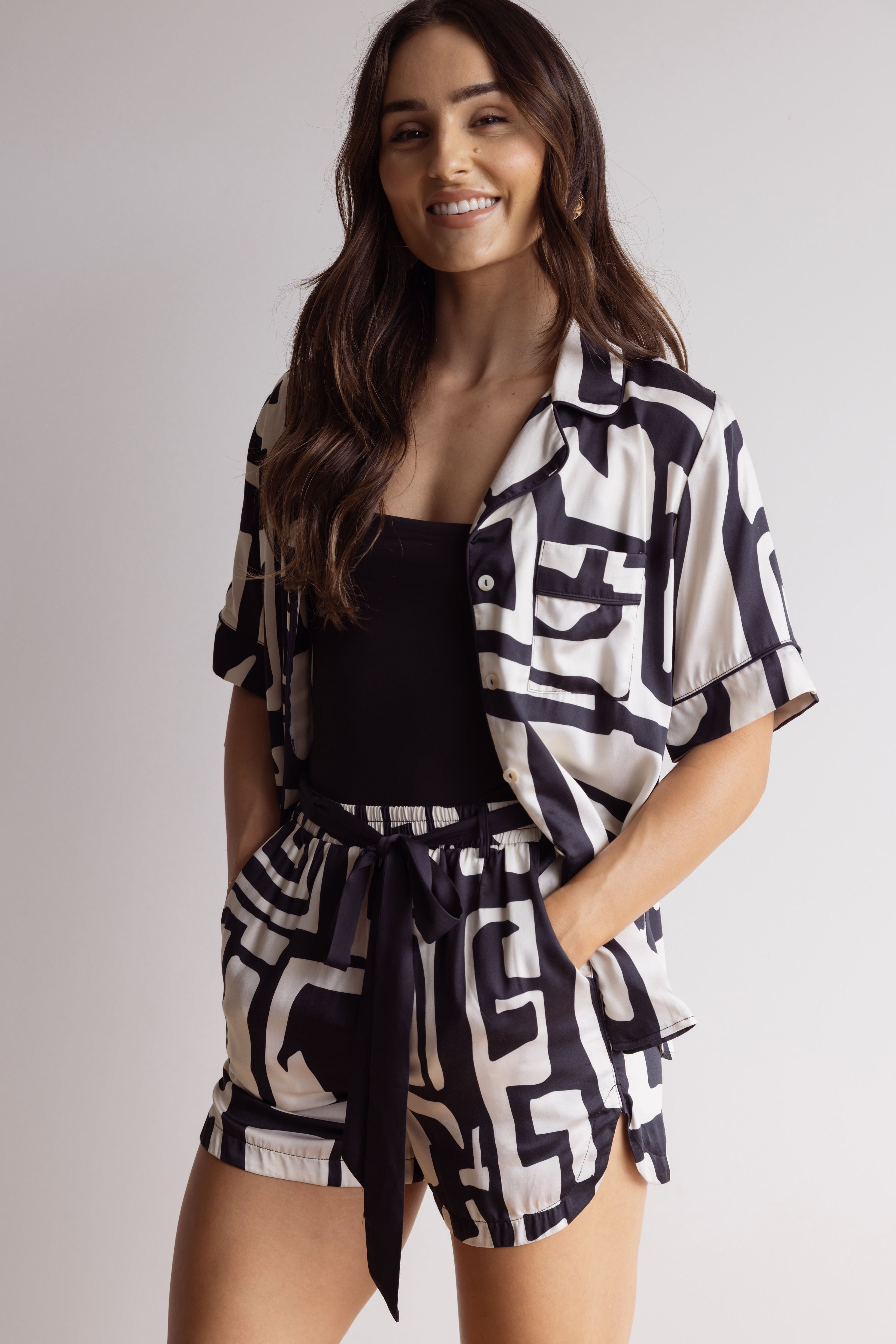 Woman smiling wearing a short bamboo PJ set with a bold black and cream geometric pattern, hands in pockets, and waistband tied in a bow.