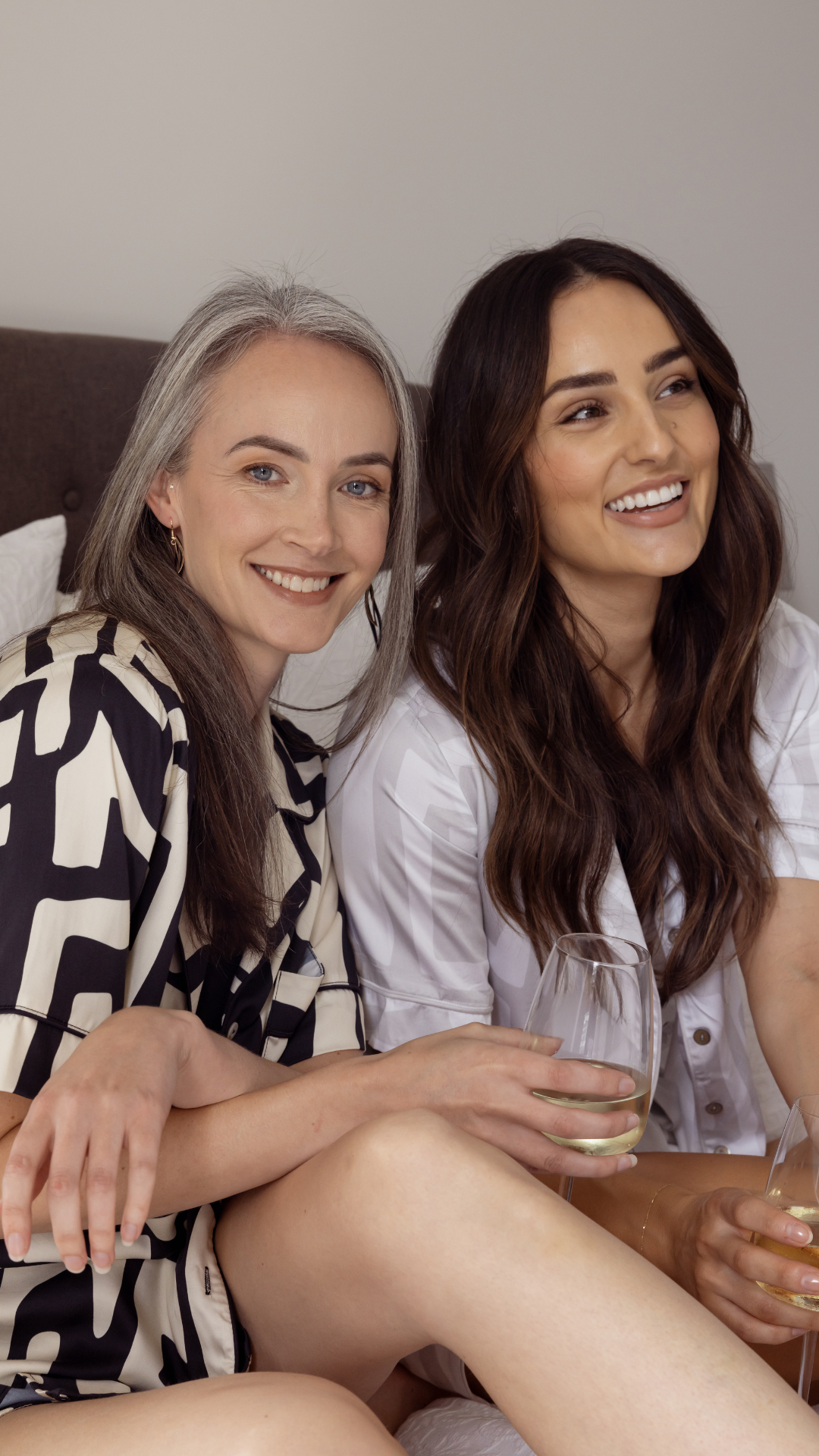 Two women sitting on a bed, smiling, in luxurious bamboo short pyjama sets with bold geometric prints—one in light grey and white, the other in black and cream