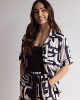 Woman wearing comfortable, breathable 100% bamboo PJ set showing a short sleeve pyjama top with button up design, piping, shorts with pockets and elasticated waistband. All in a bold geometric print in black and cream.
