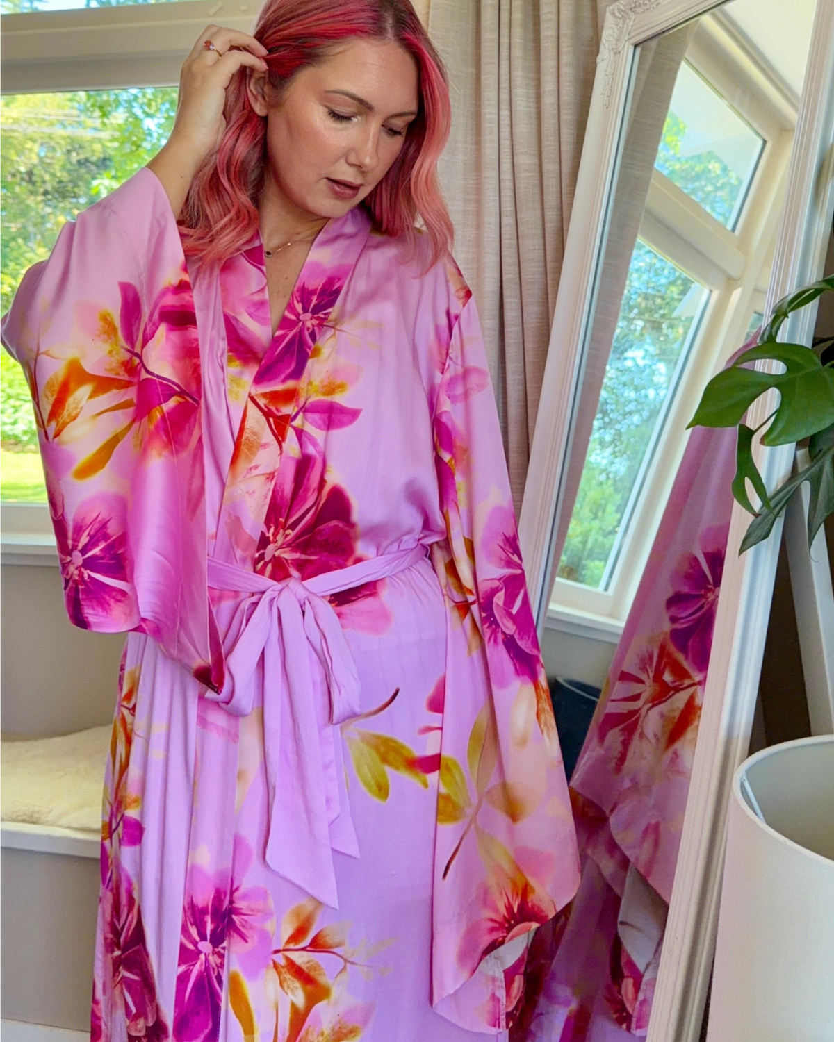 Woman wearing a bold pink floral kimono robe, with dramatic wide sleeves, waist tie and made from silky soft bamboo satin.