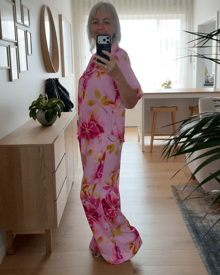Woman wearing a pink floral pyjama set taking a mirror selfie in a room with wooden flooring and furniture.