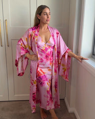 Woman wearing a pink floral kimono and matching slip dress with a low neckline, standing by a window.