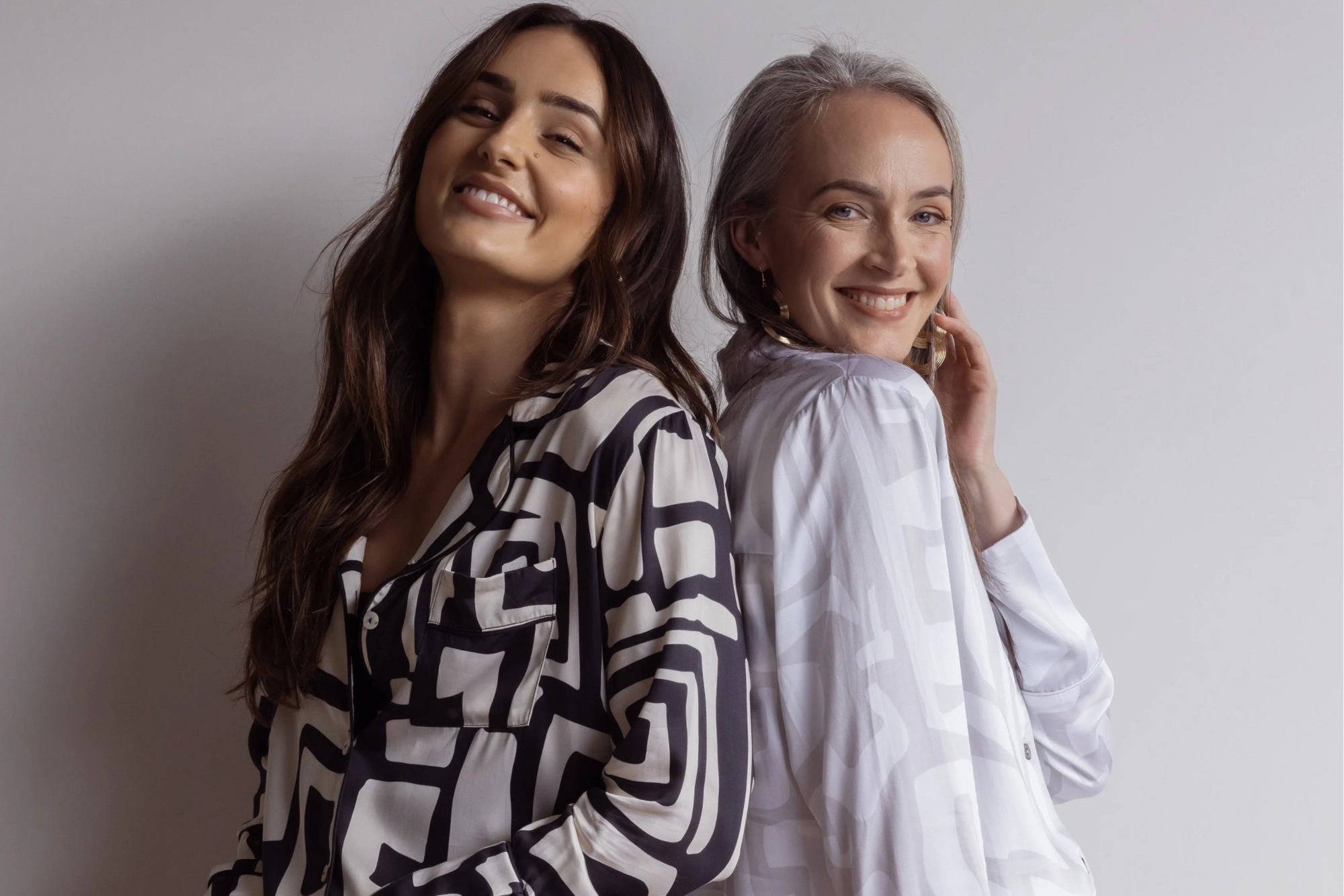 Two women standing back to back smiling, wearing beautiful bamboo pj sets with a bold geometric pattern, one in black and cream, the other in white and grey colours.