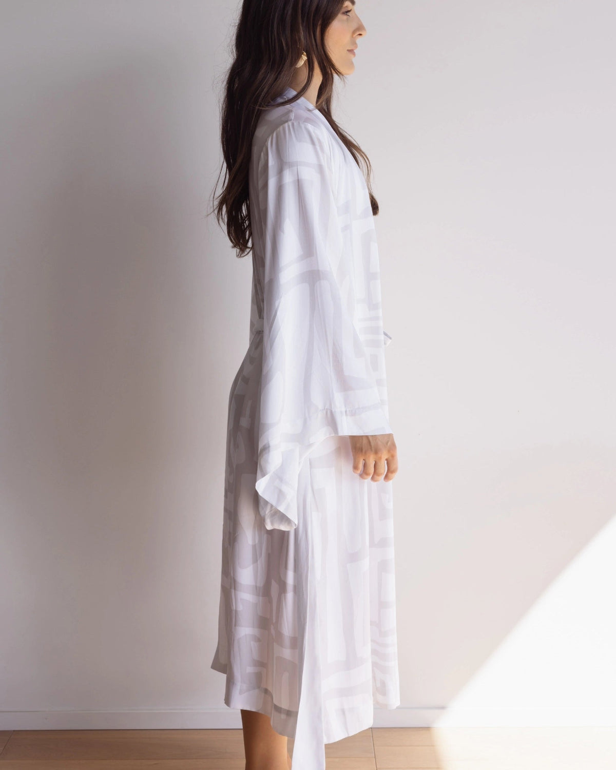 Woman wearing a comfortable kimono-style robe with wide sleeves. Made from silky-soft, breathable bamboo. Featuring a unique print design in light grey and white, with and a tie waist for an adjustable fit and hidden pockets. Perfect for relaxing.