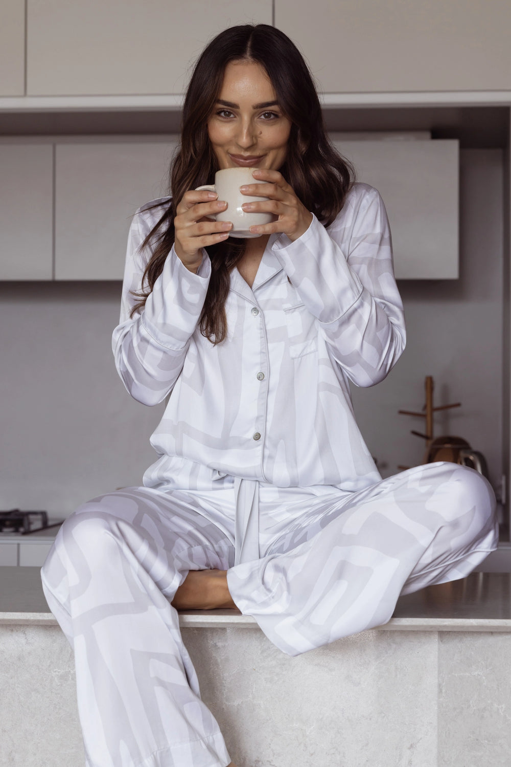 Woman in white and grey pyjamas holding a mug sitting in a kitchen.