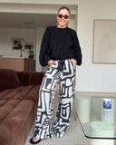 Customer wearing bamboo satin pyjama pants in bold geometric pattern, paired with a black jumper and thongs.