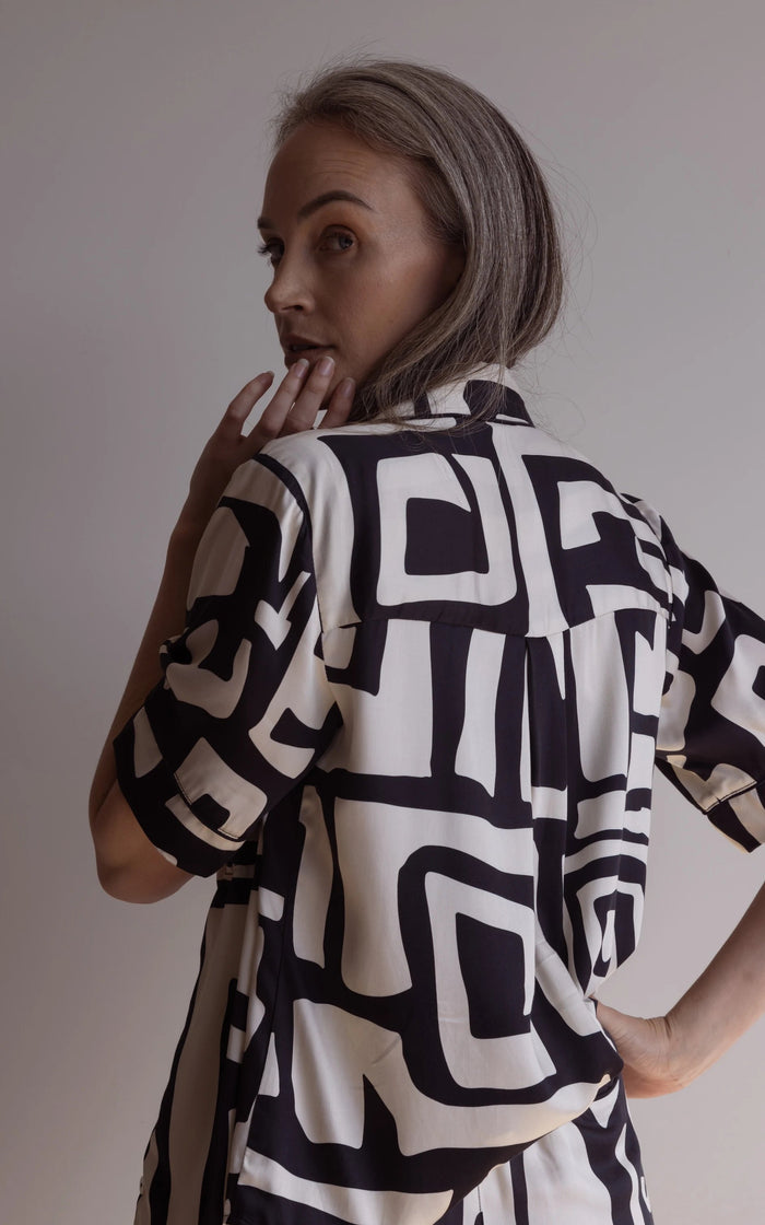 Woman looking over her shoulder wearing a short sleeve pyjama top and pants made of silky bamboo, with a bold geometric print in black and cream colour.