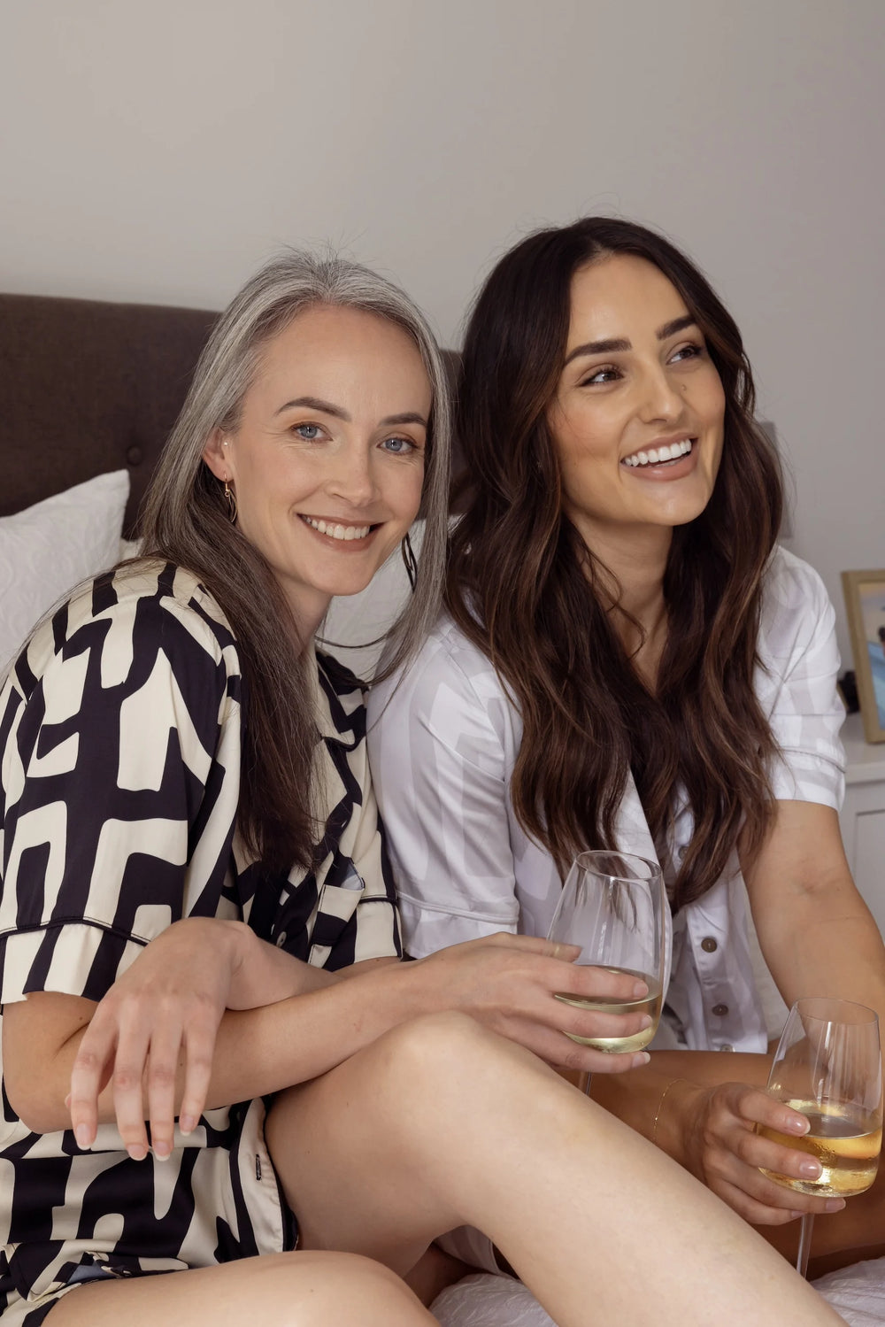 Two women sitting on a bed holding glasses of wine, smiling. Wearing matching satin bamboo pyjamas.