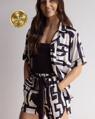 A woman modeling a matching black and white geometric print short-sleeve button-up shirt and tie-waist shorts pyjama set, paired with a black tank top. A gold 'BEST SELLER' badge is visible in the top left corner.
