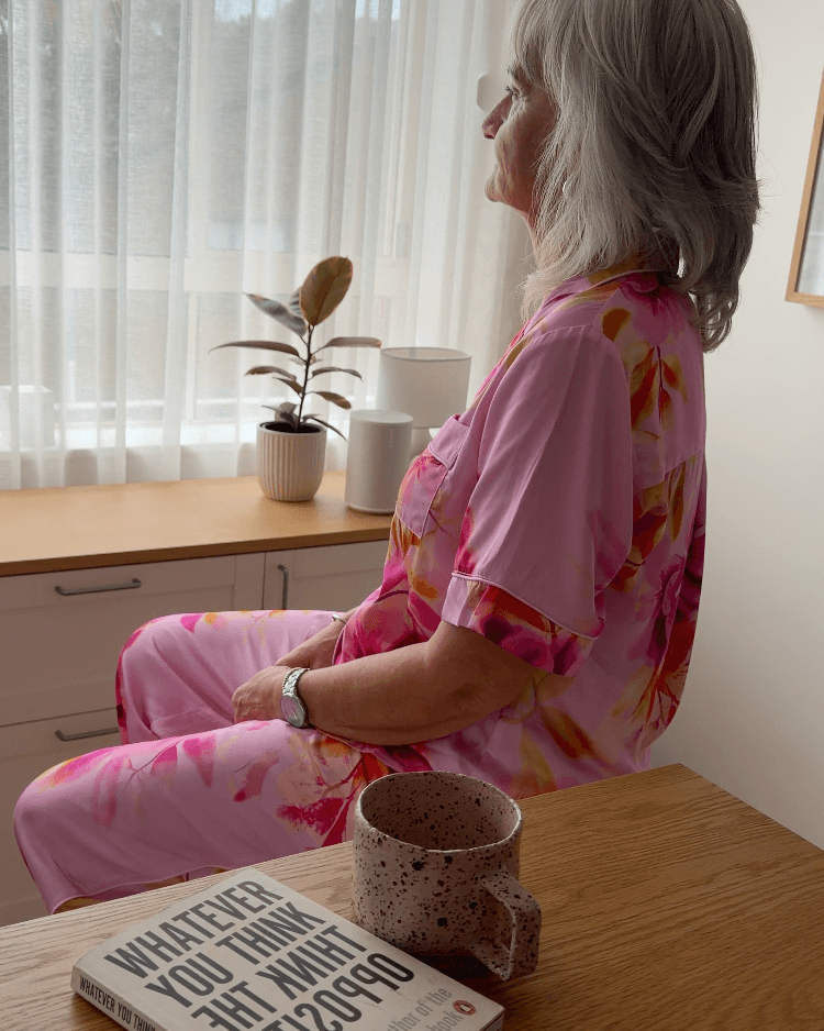 Woman in pink floral pajamas sitting at a table with a book and mug, looking out a window.