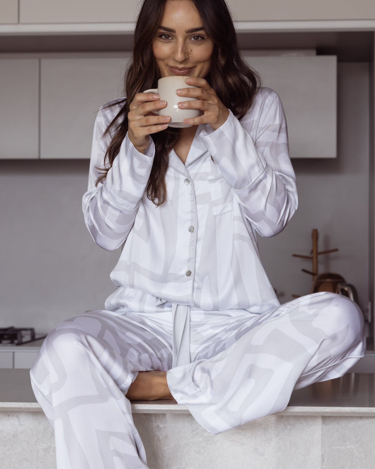 Woman wearing a long sleeved shirt and matching pants. Pyjama set made from silky soft bamboo satin. Whilst sitting on a kitchen bench drinking coffee.