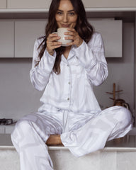 Woman wearing a long sleeved shirt and matching pants. Pyjama set made from silky soft bamboo satin. Whilst sitting on a kitchen bench drinking coffee.
